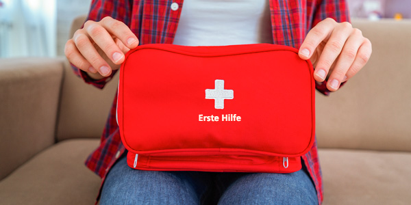 Person sitzt auf beigem Sofa und hält mit beiden Händen einen roten Erste-Hilfe-Kasten mit weißem Kreuz und der Aufschrift „Erste Hilfe“. Kleidung: blaue Jeans und rot karierte Bluse.
